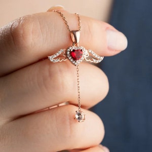 May include: A rose gold necklace with a red heart pendant and clear crystal wings. The pendant hangs from a delicate chain with a single clear crystal at the end.