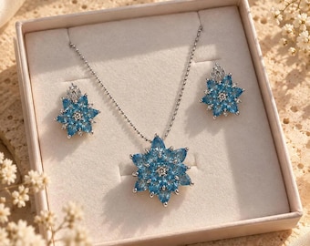 May include: A jewelry set featuring a silver-toned necklace and matching stud earrings, each designed with a central cluster of marquise-cut light blue stones arranged in a starburst pattern, presented in a square, light-colored gift box.