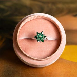 May include: A silver ring with a starburst design featuring multiple green gemstones and a central clear stone. The ring is presented in a pink velvet ring box, set against a warm, textured background.