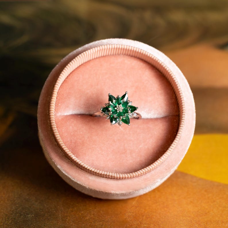 May include: A silver ring with a starburst design featuring multiple green gemstones and a central clear stone. The ring is presented in a pink velvet ring box, set against a warm, textured background.
