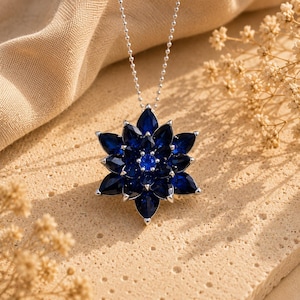 Peut inclure: Un collier argenté avec un pendentif en forme de fleur à plusieurs couches. Le pendentif est orné de nombreuses pierres bleu foncé taillées en marquise disposées en cercles concentriques, avec une pierre bleue ronde au centre.