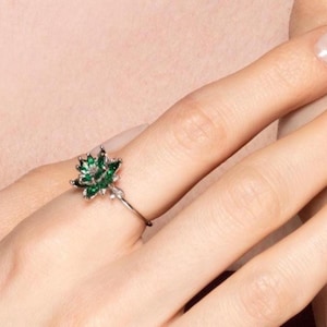 May include: A silver ring featuring a detailed green gemstone flower design. The ring's floral centerpiece is composed of multiple green stones, creating a vibrant and eye-catching accessory. The ring is worn on a finger.