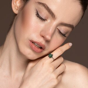 May include: A silver ring with a flower-shaped design featuring multiple small, dark green gemstones. The ring is worn on a finger, with the person's face and shoulder in the background. The person has their eyes closed.