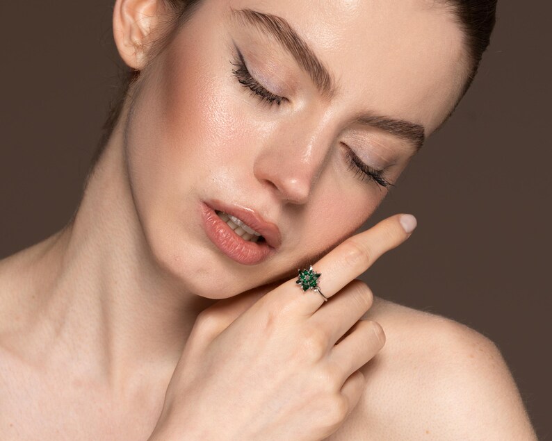 May include: A silver ring with a flower-shaped design featuring multiple small, dark green gemstones. The ring is worn on a finger, with the person's face and shoulder in the background. The person has their eyes closed.