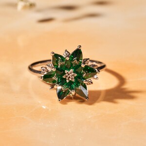 May include: A silver ring featuring a flower design with green gemstone petals and a small, central cluster. The ring is set against a warm, neutral background, casting a shadow.