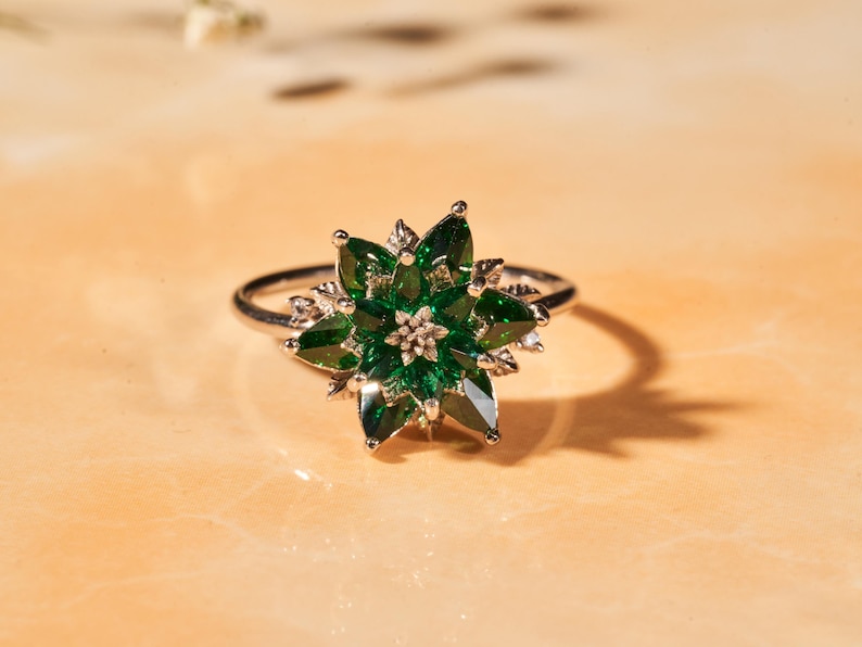 May include: A silver ring featuring a flower design with green gemstone petals and a small, central cluster. The ring is set against a warm, neutral background, casting a shadow.