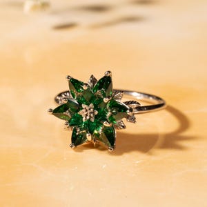 May include: A silver ring featuring a flower design with emerald green gemstone petals. The ring's center has a cluster of smaller gemstones. The ring is set against a light-colored background.