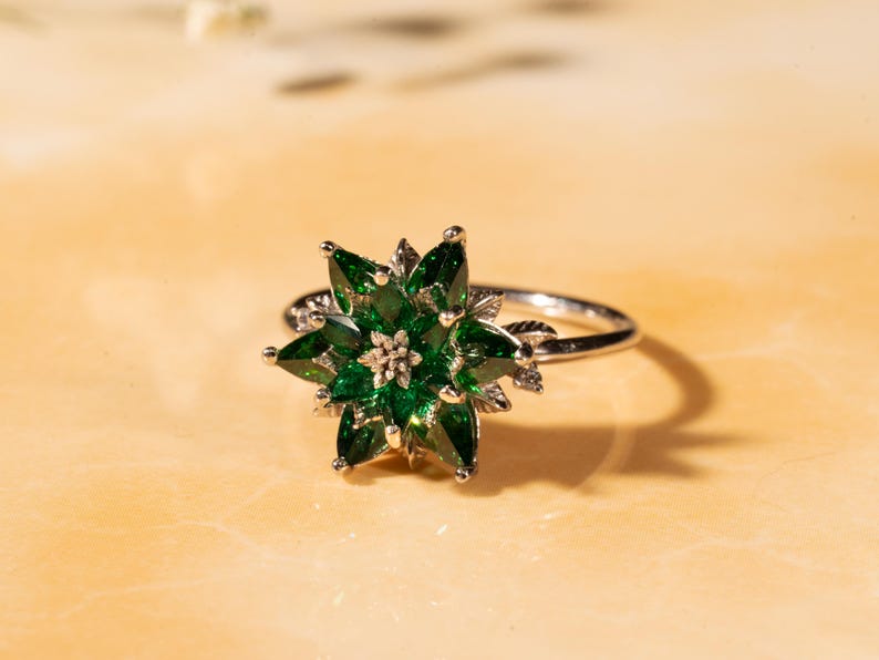 May include: A silver ring featuring a flower design with emerald green gemstone petals. The ring's center has a cluster of smaller gemstones. The ring is set against a light-colored background.