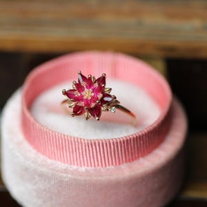 Op de afbeelding: Een gouden ring met een bloemmotief, gezet met robijnkleurige edelstenen. De ring wordt gepresenteerd in een roze fluwelen ringdoosje met een roze lint. Close-up, met de nadruk op de fijne details van het bloemendesign.