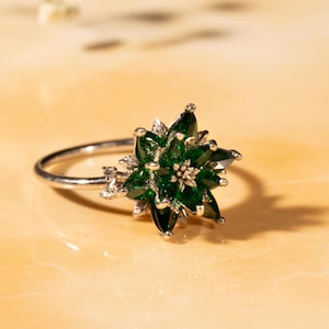 May include: A silver ring featuring a floral design with emerald green gemstones. The ring's center is a cluster of smaller stones, and it has a delicate band. The ring is set against a light-colored background.