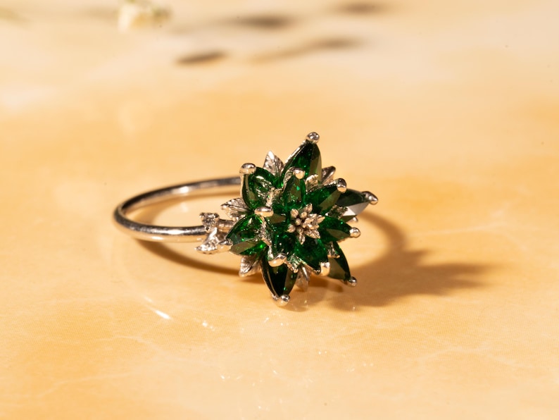 May include: A silver ring featuring a floral design with emerald green gemstones. The ring's center is a cluster of smaller stones, and it has a delicate band. The ring is set against a light-colored background.