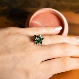 May include: A silver ring featuring a flower design with dark green gemstones and small clear stones. The ring is set on a hand, with a blurred background.