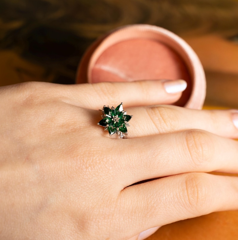 May include: A silver ring featuring a flower design with dark green gemstones and small clear stones. The ring is set on a hand, with a blurred background.