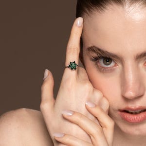 May include: A close-up of a ring with a dark band and a star-shaped cluster of green gemstones. The ring is on a person's finger, with the person's face in the background. The ring's design is intricate and eye-catching.