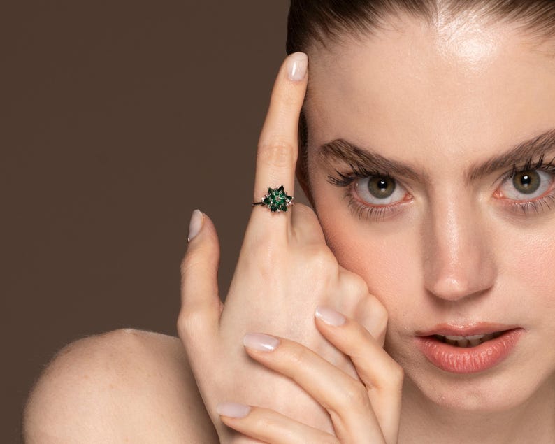May include: A close-up of a ring with a dark band and a star-shaped cluster of green gemstones. The ring is on a person's finger, with the person's face in the background. The ring's design is intricate and eye-catching.