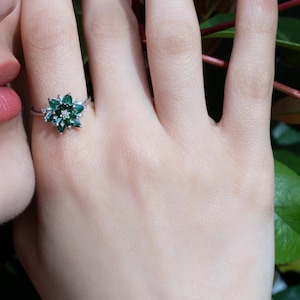 May include: A silver ring featuring a floral design with emerald-green gemstones and a central diamond. The ring is worn on a hand with a light skin tone, with a blurred background of greenery.