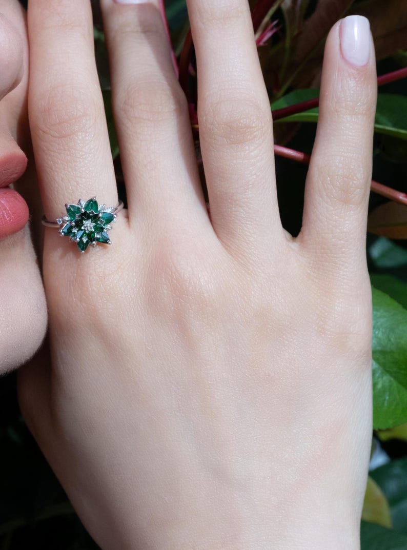 May include: A silver ring featuring a floral design with emerald-green gemstones and a central diamond. The ring is worn on a hand with a light skin tone, with a blurred background of greenery.