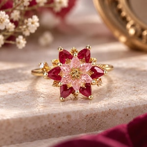 May include: A gold ring featuring a floral design with a centre of light pink pear-shaped stones surrounded by a layer of deep red pear-shaped stones. The metal band is thin and polished, with small gold leaf-like accents between the petals.
