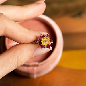 May include: A floral ring with a flower design. The ring features a central cluster of yellow gemstones surrounded by red gemstones, creating a vibrant contrast. The ring is held in a pink velvet jewelry box.