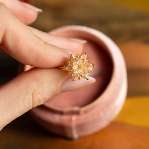 May include: A gold-colored ring with a floral design, featuring multiple light pink gemstones. The ring is held in front of a pink velvet ring box. The ring's design resembles a star or flower.