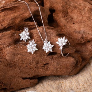 May include: A jewellery set consisting of a necklace, two earrings, and a ring, all featuring a silver-toned metal base and white, petal-shaped stones arranged in a floral pattern. The necklace includes a delicate silver-toned ball chain.