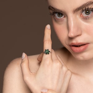 May include: A silver ring featuring a flower-shaped design with dark green gemstones. The ring is set against a neutral background, showcasing the jewelry's intricate details and color.