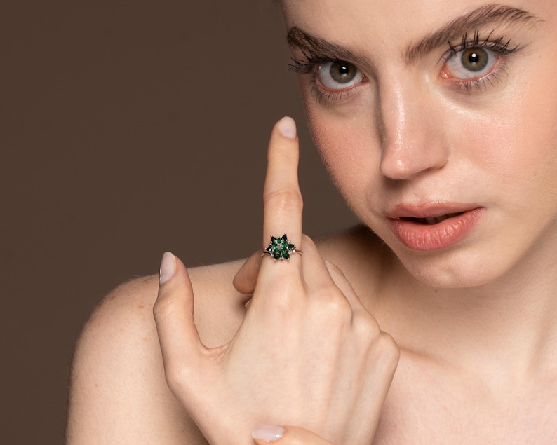 May include: A silver ring featuring a flower-shaped design with dark green gemstones. The ring is set against a neutral background, showcasing the jewelry's intricate details and color.