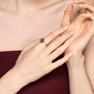May include: A silver ring featuring a cluster of small, dark green gemstones in a floral design. The ring is worn on a finger, with the hand and arm visible against a burgundy background.