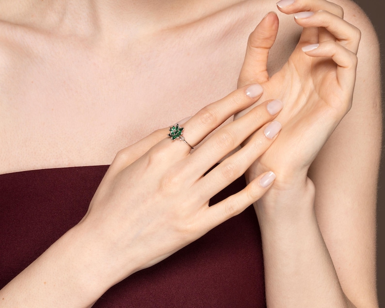 May include: A silver ring featuring a cluster of small, dark green gemstones in a floral design. The ring is worn on a finger, with the hand and arm visible against a burgundy background.