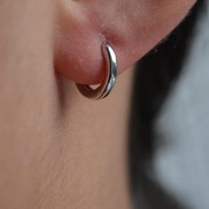Peut inclure: Une petite boucle d'oreille argentée est portée dans un piercing à l'oreille. La boucle d'oreille a un design simple et arrondi, reflétant la lumière. La peau autour de l'oreille est visible, avec un arrière-plan flou.