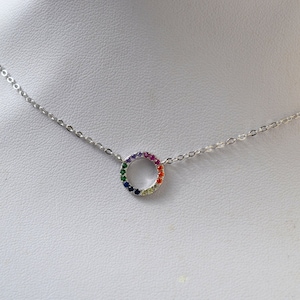 Peut inclure: Un collier en chaîne argent avec un petit pendentif rond. Le cercle est serti de pierres précieuses colorées dans un motif arc-en-ciel.