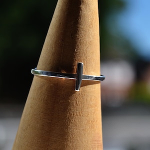 Op de afbeelding: Een zilverkleurige ring met een eenvoudig kruisontwerp. De ring wordt getoond op een houten object, waarbij het gepolijste oppervlak en de minimalistische stijl worden benadrukt. Het kruis is horizontaal geplaatst.