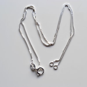 Op de afbeelding: Een zilveren ketting met een box chain design. De ketting heeft een karabijnsluiting en twee kleine sprongringen. De ketting is gemaakt van kleine, vierkante schakels en wordt gepresenteerd op een wit oppervlak.