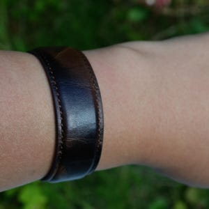 Peut inclure: Un bracelet en cuir marron foncé avec un bord cousu. Le bracelet est porté sur un bras, mettant en valeur son design simple et classique. Le cuir semble légèrement brillant, suggérant une finition polie. La largeur du bracelet est d'environ 2,5 cm.
