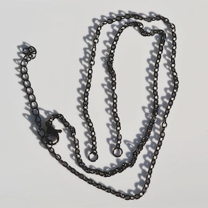 May include: A dark gunmetal-coloured chain necklace featuring oval-shaped links. The necklace includes a lobster claw clasp and an adjustable extender chain with a small circular charm at the end, creating a simple and functional jewellery piece.