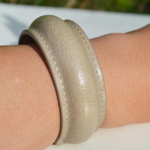 May include: A beige leather bracelet with a rounded design. The bracelet is worn on an arm and has a stitched seam detail. The bracelet's colour is a neutral tone, suitable for various outfits.
