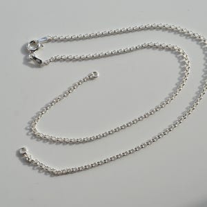 May include: A delicate silver chain necklace with a lobster clasp closure. The chain is composed of small, round links. The necklace is displayed on a white surface, with shadows adding depth.
