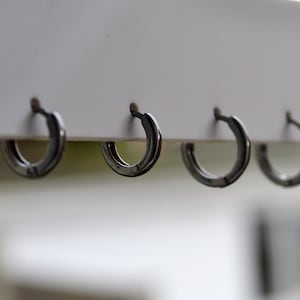 Puede incluir: Cuatro pequeños pendientes de aro de color plateado se muestran sobre una superficie blanca. Los pendientes están espaciados uniformemente y tienen un diseño simple y minimalista. El fondo está borroso.