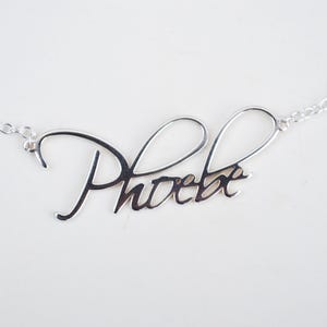 Peut inclure: Un collier argenté avec le prénom "Phoebe" écrit en cursive. Le nom est l'élément central, relié à une fine chaîne de chaque côté. Le collier est présenté sur un fond blanc uni.