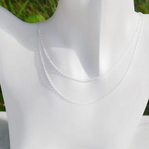 May include: A double-layered silver chain necklace featuring delicate, small circular links. The necklace is displayed on a white mannequin bust, showcasing the reflective metallic finish and the two distinct strands that drape gracefully around the neck area.