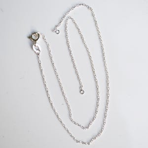 May include: A delicate silver chain necklace with a lobster clasp closure. The chain is composed of small, oval-shaped links. The necklace is displayed on a white surface. The chain is approximately 45 cm long, showcasing its simple elegance.