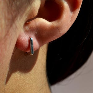 May include: A close-up of a silver rectangular earring. The earring is a small, minimalist design, and is worn on the ear. The earring is a simple, modern piece of jewellery.
