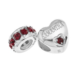 Puede incluir: Dos abalorios de plata. Uno tiene forma de corazón con el nombre "SAMANTHA" grabado y una pequeña gema roja. El otro es un cilindro plateado con múltiples gemas rojas.