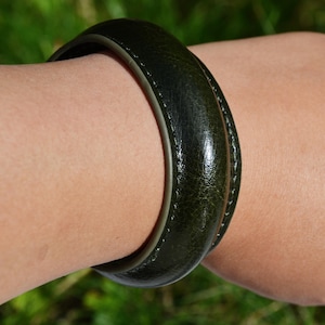 May include: A dark green leather bangle. The circular bracelet has a textured surface and is worn on a wrist. The bracelet's design is simple and elegant. The circumference is approximately 20 cm.