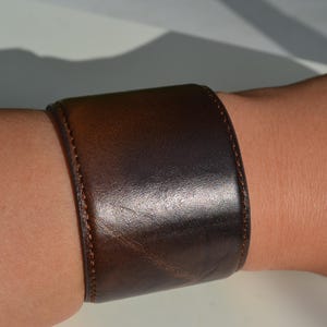Op de afbeelding: Een brede, donkerbruine leren manchetarmband. De armband is gestikt met een lichtere bruine draad en lijkt om een pols te worden gedragen. Het leer heeft een lichte glans, wat suggereert dat het gepolijst kan zijn.