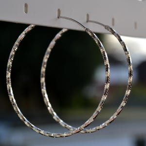 May include: A pair of large, silver hoop earrings. The hoops have a textured, mosaic-like pattern, reflecting light. The earrings are suspended from a white display board, with a blurred background. The earrings are a classic jewelry item.