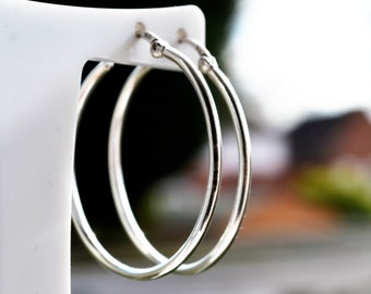 May include: A pair of silver hoop earrings. The earrings are circular and have a polished finish. They are presented on a white display stand. The background is blurred, with green and brown tones.