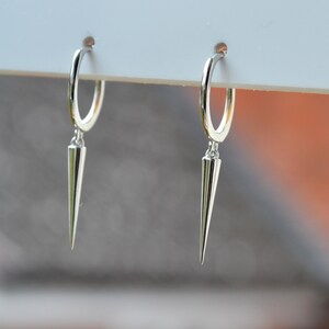 May include: A pair of silver hoop earrings with a dangling, pointed cone charm. The earrings are made of a shiny, reflective metal and hang from a white surface. The background is blurred, with hints of orange and gray.