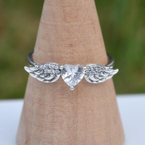 May include: A silver ring with a clear, heart-shaped gemstone as the focal point, set between detailed wing motifs. The wings are embellished with small, clear stones. The ring is presented on a wooden display stand.