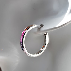 Può includere: Un orecchino a cerchio d'argento con una fila di pietre preziose colorate. Le pietre preziose sono disposte in un motivo arcobaleno, che passa dal blu al rosso. L'orecchino è mostrato su uno sfondo bianco.